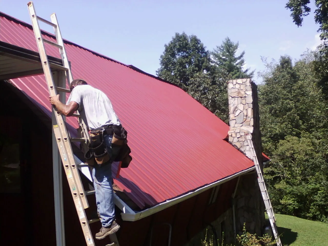 Roofer on a ladder beside a red metal roof and stone chimney — Soffit & Fascia Repair by Roofing Brothers in Northfield